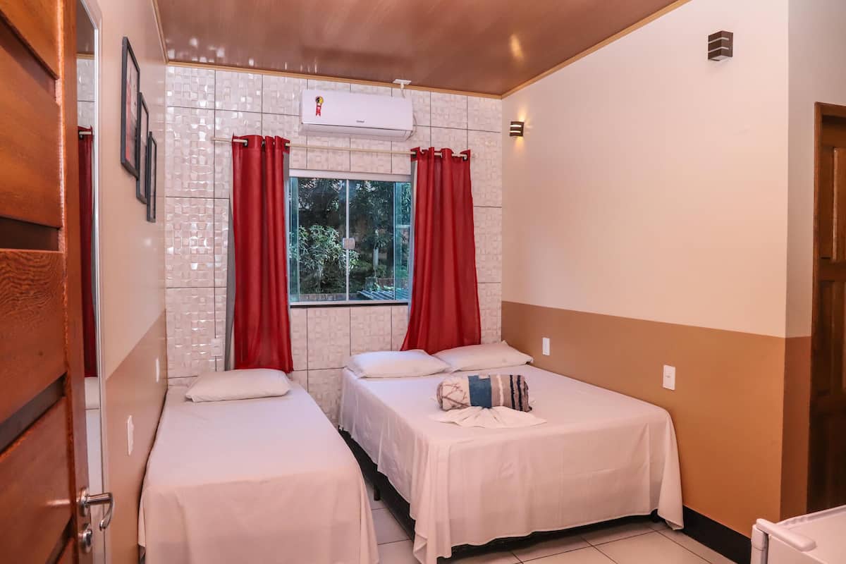 A hotel room with two beds, a window with red curtains, an air conditioning unit, and a view of greenery outside.