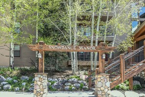A wooden sign with the text "SNOWDANCE MANOR" above a stone pillar, with a staircase leading to a building in the background. - Snow Dance Manor 301 (Keystone)