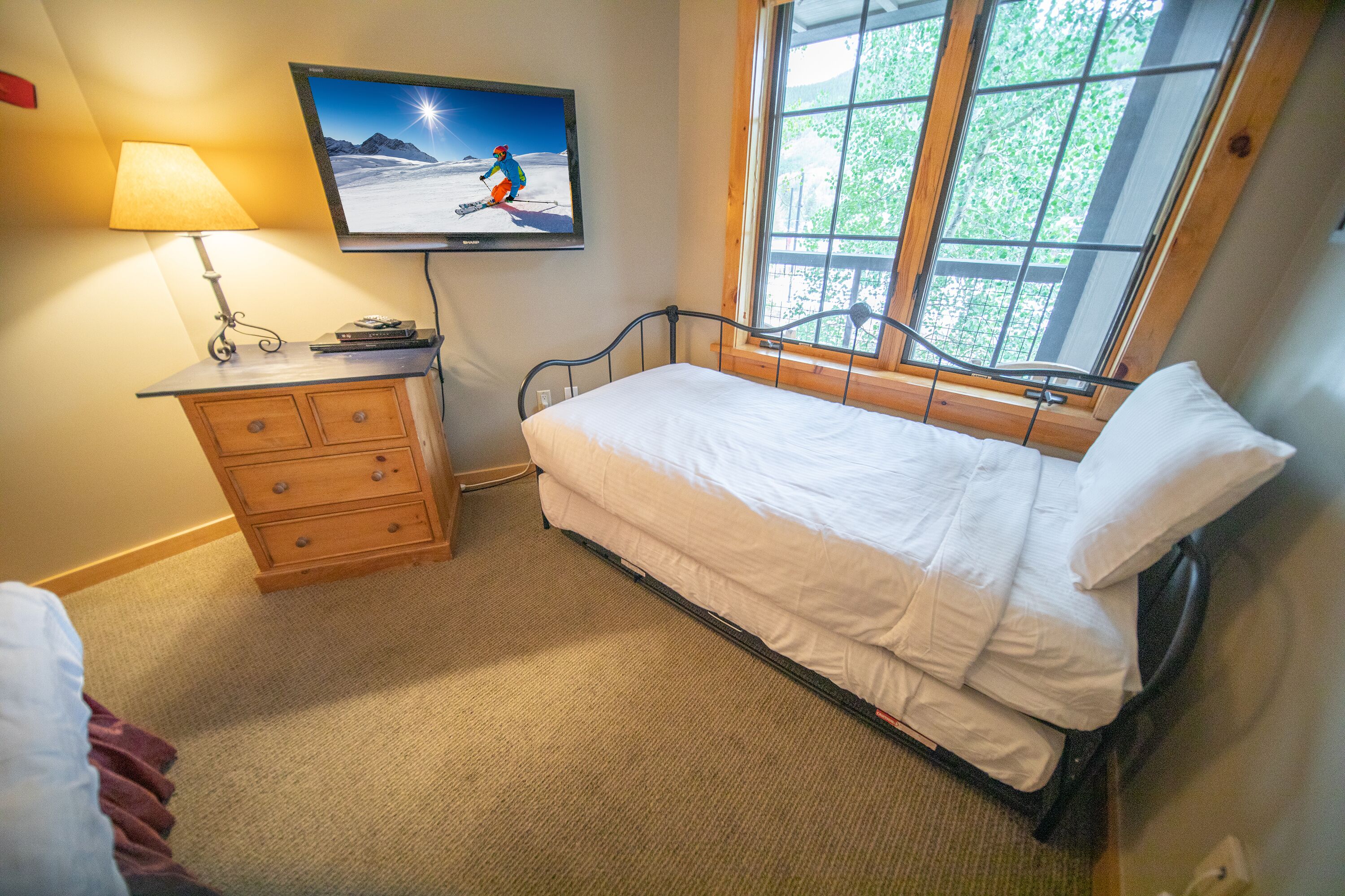Una cama pequeña y bien hecha con ropa blanca en una habitación con un aparador de madera, un televisor montado en la pared y una ventana con vistas a la vegetación.
