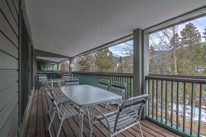 Una terraza cubierta al aire libre con una mesa y sillas, barandilla y vistas a árboles y montañas.