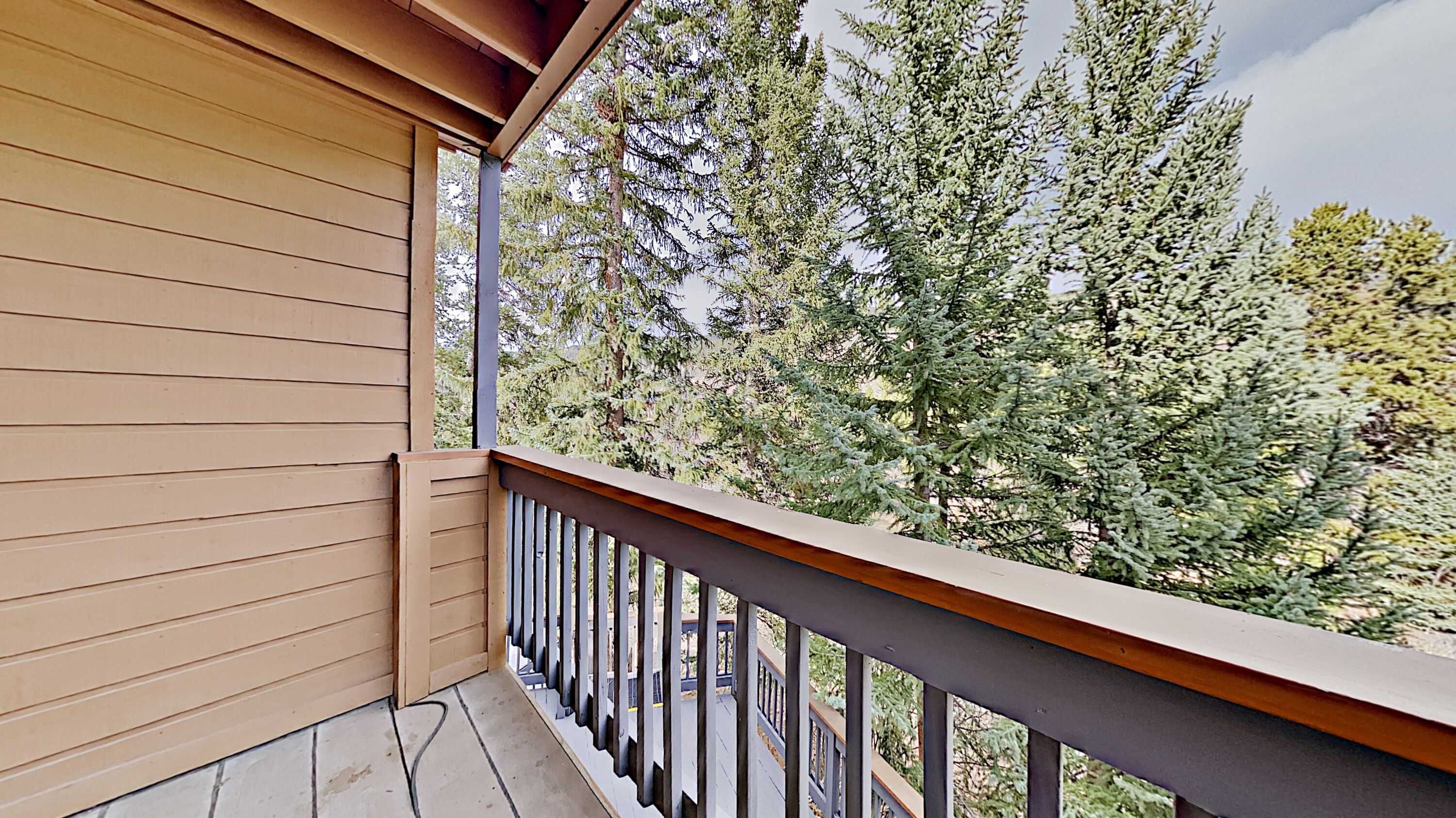 A wooden balcony with a metal railing, overlooking a dense forest of evergreen trees.