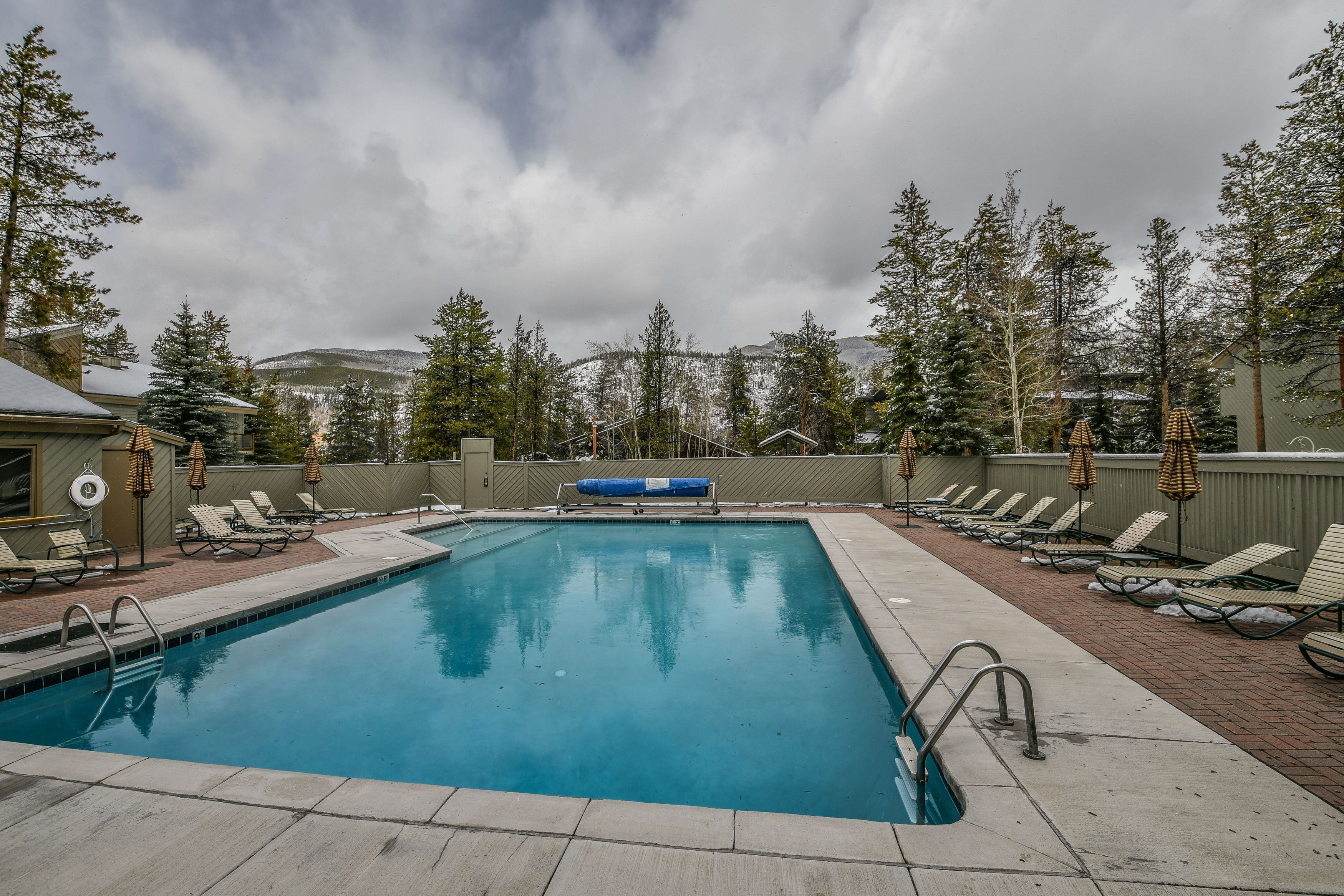 A rectangular swimming pool with a blue water surface, surrounded by lounge chairs and umbrellas, with a view of snow-covered mountains in the background.