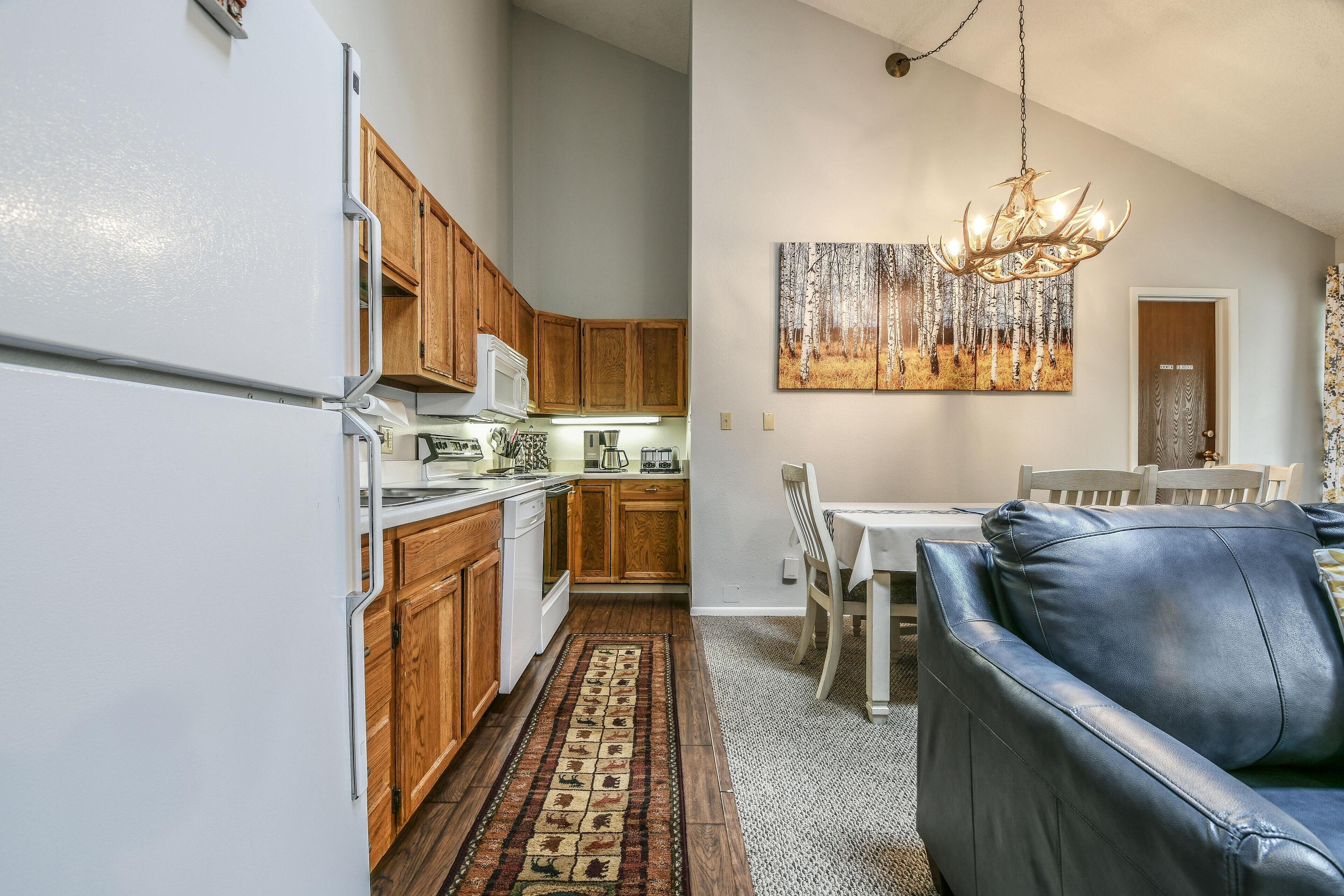 A kitchen with wooden cabinets, a white refrigerator, and a dining area with a leather couch and a chandelier.