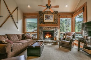 Un salón acogedor con chimenea de piedra, muebles de cuero y grandes ventanales con vistas a un bosque.