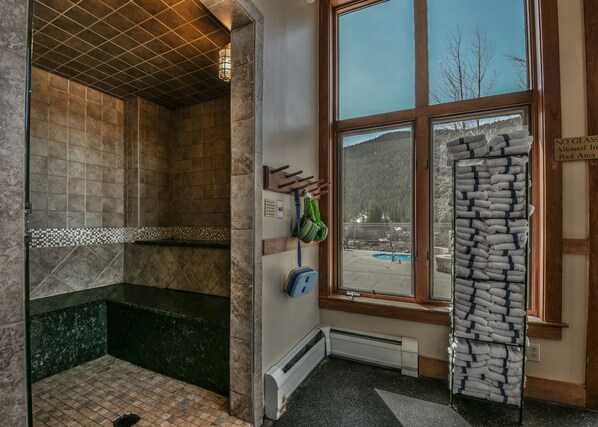 Une salle de bain avec une douche, un porte-serviettes, une fenêtre donnant sur une piscine et des arbres, et un panneau indiquant qu’il est interdit d’apporter des objets en verre dans l’espace piscine.