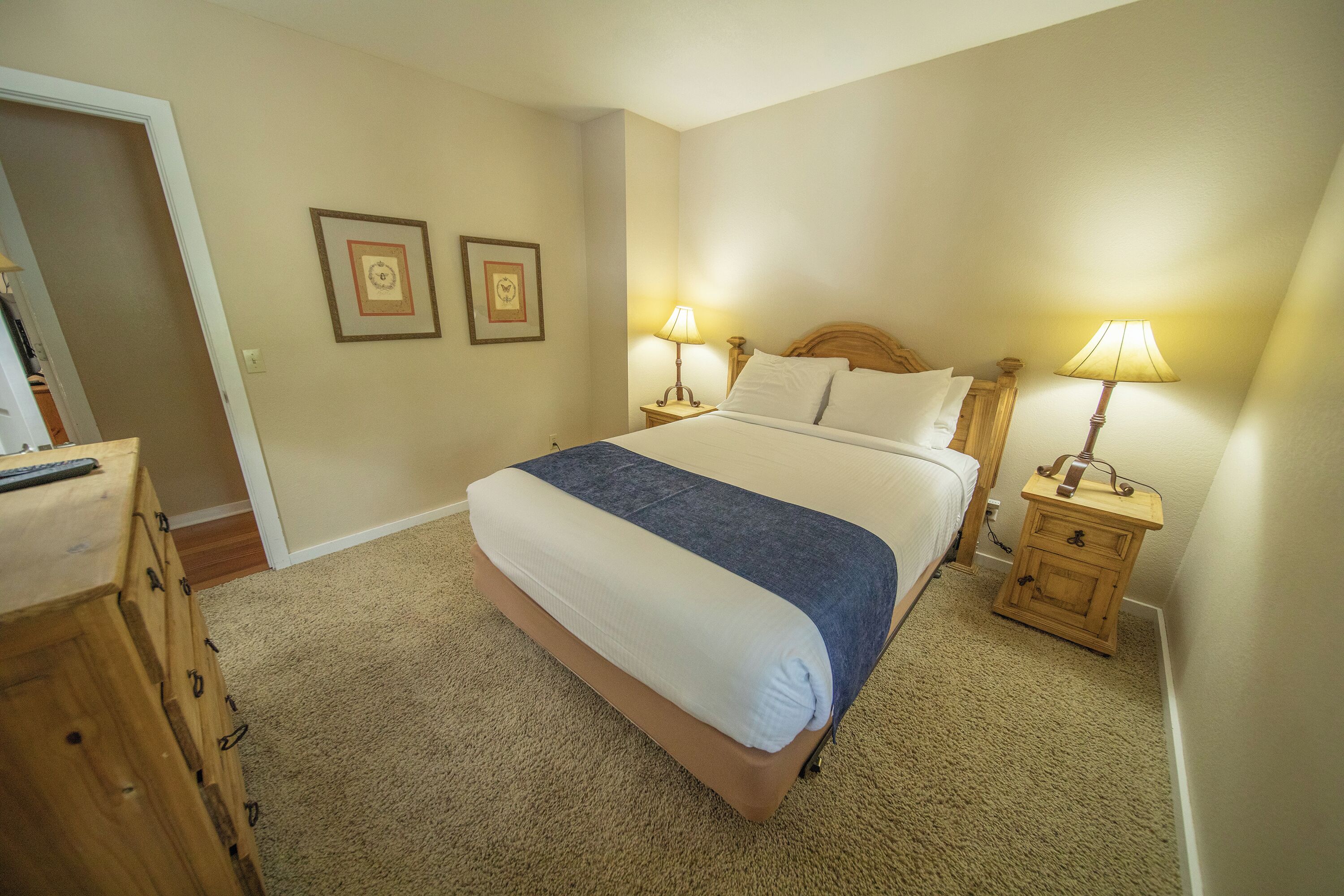 A bedroom with a large bed, two bedside tables with lamps, a wooden dresser, and two framed pictures on the wall.
