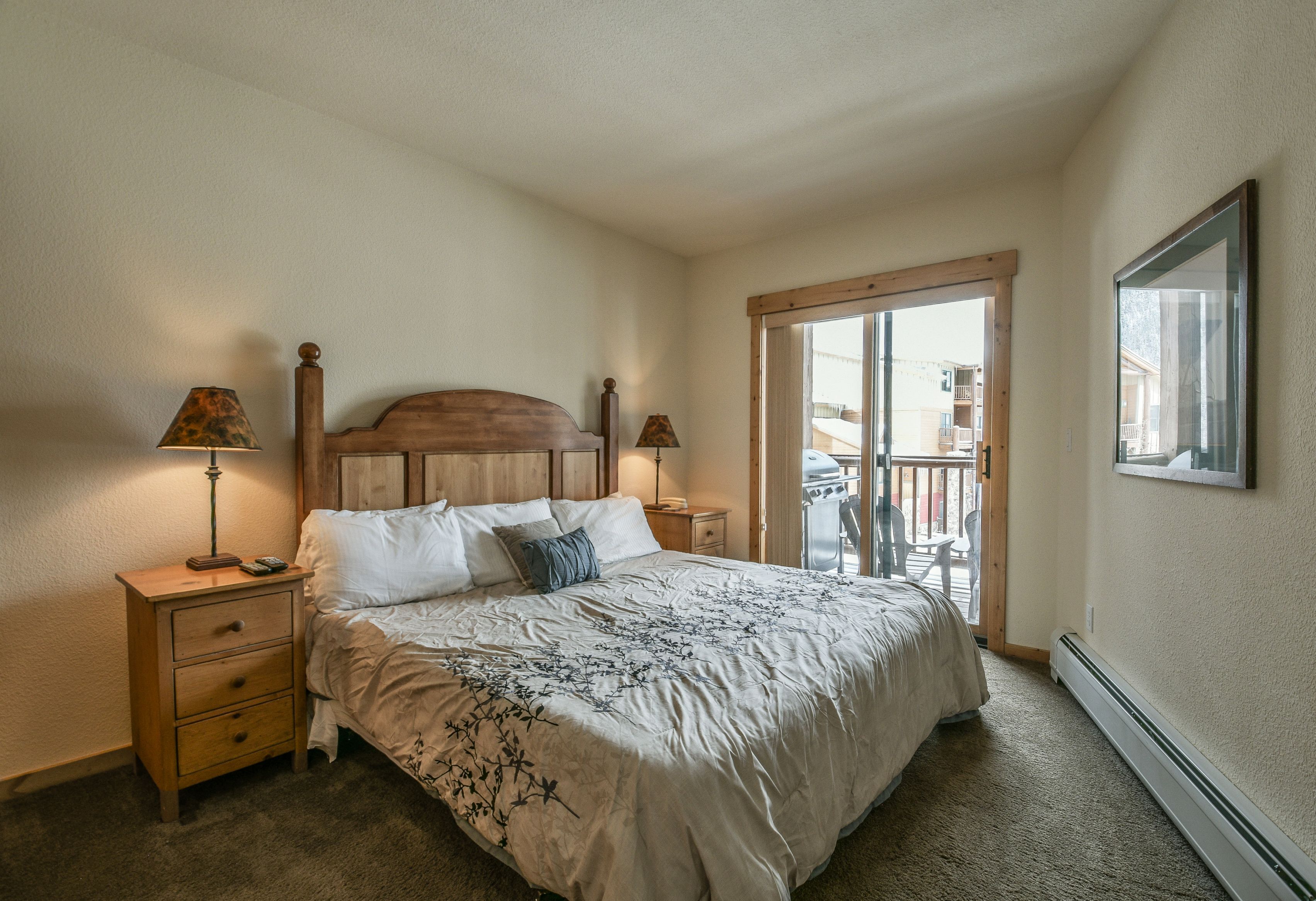 A bedroom with a wooden headboard, a bed with white bedding, two bedside tables with lamps, a sliding glass door to a balcony, and a framed picture on the wall.