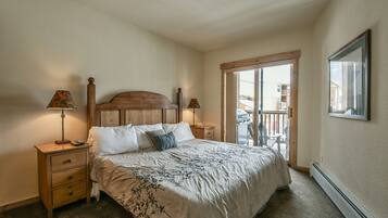 A bedroom with a wooden headboard, a bed with white bedding, two bedside tables with lamps, a sliding glass door to a balcony, and a framed picture on the wall.