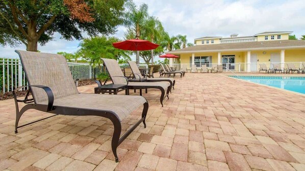 Un espace aménagé au bord de la piscine, avec des chaises longues et des parasols, une terrasse en briques et un bâtiment en arrière-plan.