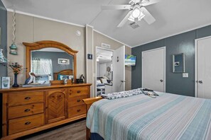 Une chambre à coucher comprenant un lit, une commode, un miroir, un ventilateur de plafond et un téléviseur fixé au mur.