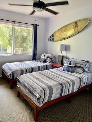 A bedroom with two beds, a ceiling fan, a surfboard on the wall, and a window with curtains.