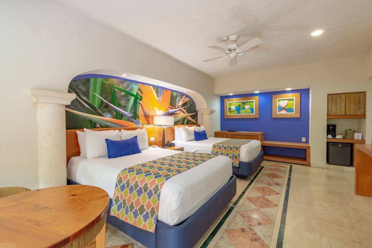 Une chambre d’hôtel avec deux lits, une petite table, un ventilateur de plafond et des œuvres d’art accrochées aux murs.