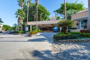 Entrada de un hotel con un letrero que dice "Beach Club & Hotel", rodeado de palmeras y un jardín paisajístico.