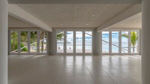 Een ruime kamer met grote ramen, met uitzicht op een strand en palmbomen.