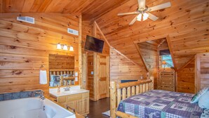 Ein gemütliches Schlafzimmer in einer Blockhütte mit einem Holzbett, einem großen Fenster, einem Deckenventilator und einem an der Wand befestigten Fernseher.
