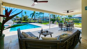 A covered outdoor area with a pool, seating, and a view of palm trees and houses.