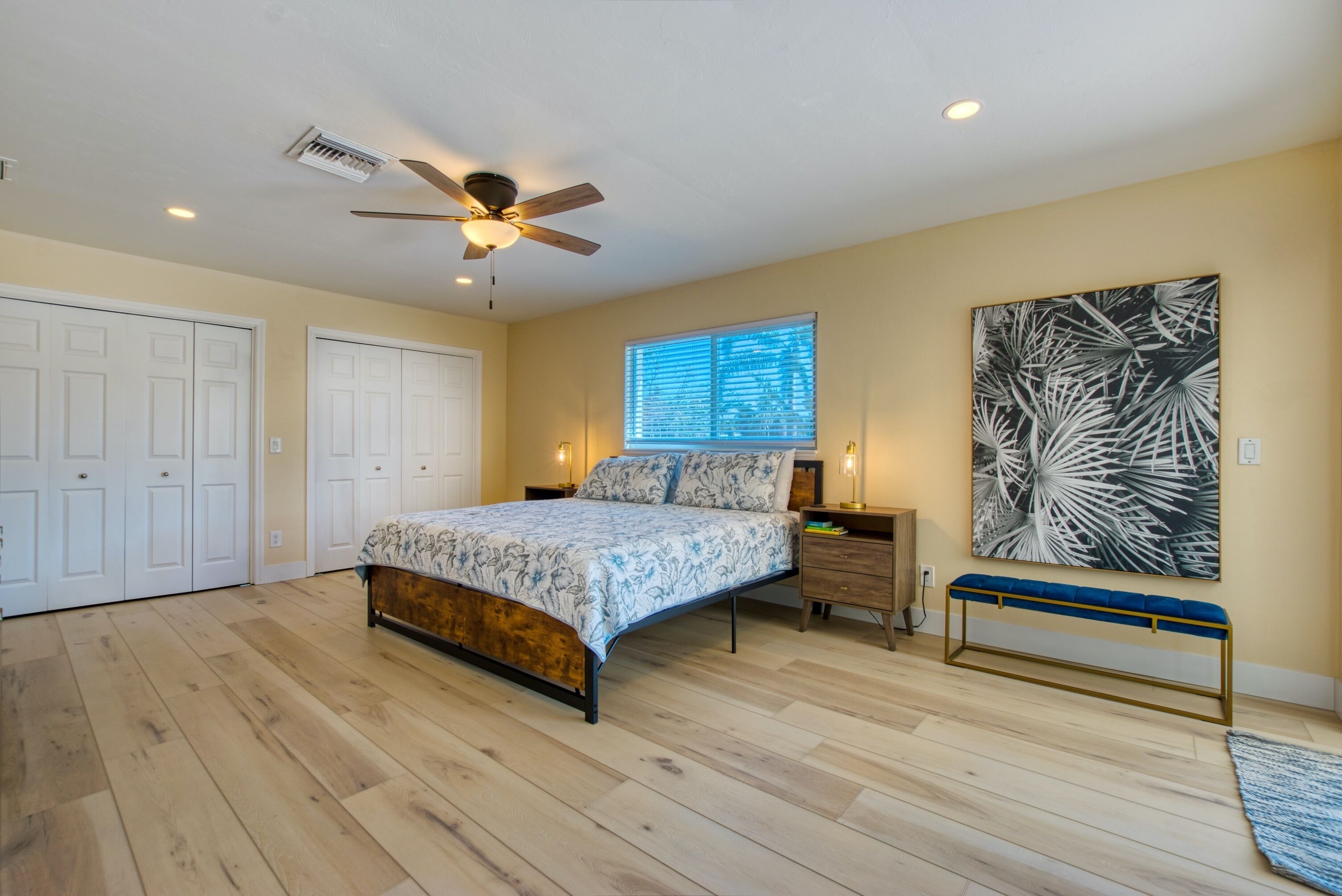 A bedroom with a bed, a ceiling fan, a window with blinds, a wall-mounted artwork, and a bench.