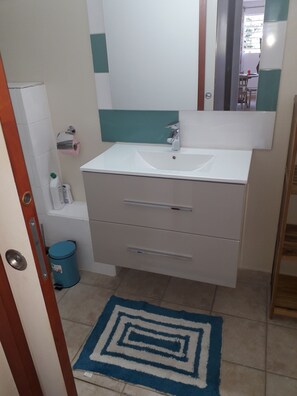 A bathroom with a white sink, a mirror, and a blue trash bin. - Gîte MORINGA (Bouillante)