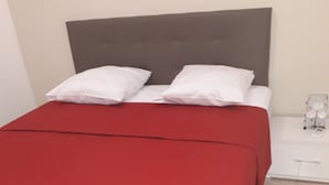 A bed with a red bedspread and white pillows, a nightstand with a clock, and a tiled floor. - Gîte MORINGA (Bouillante)