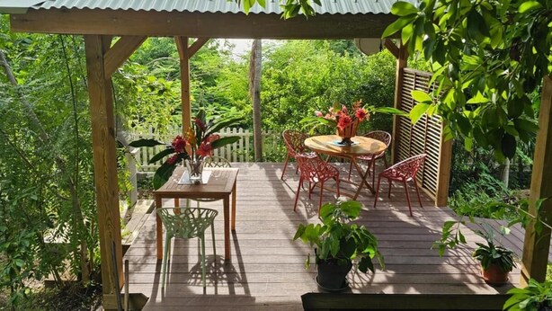 A wooden deck with a metal roof, surrounded by lush greenery. - Gîte MORINGA (Bouillante)