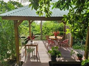 A wooden deck with a metal roof, surrounded by lush greenery. - Gîte MORINGA (Bouillante)