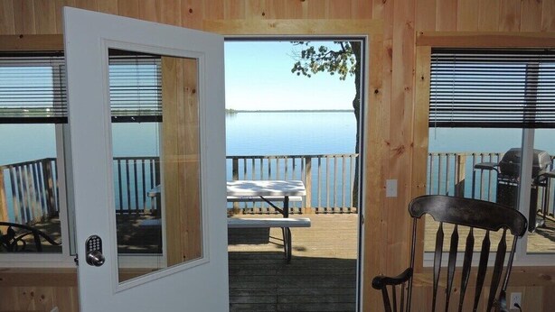 A wooden cabin with a door leading to a deck with a picnic table and a chair. - Cottage #22 - 3 bedroom/1 bath (Cape Vincent)
