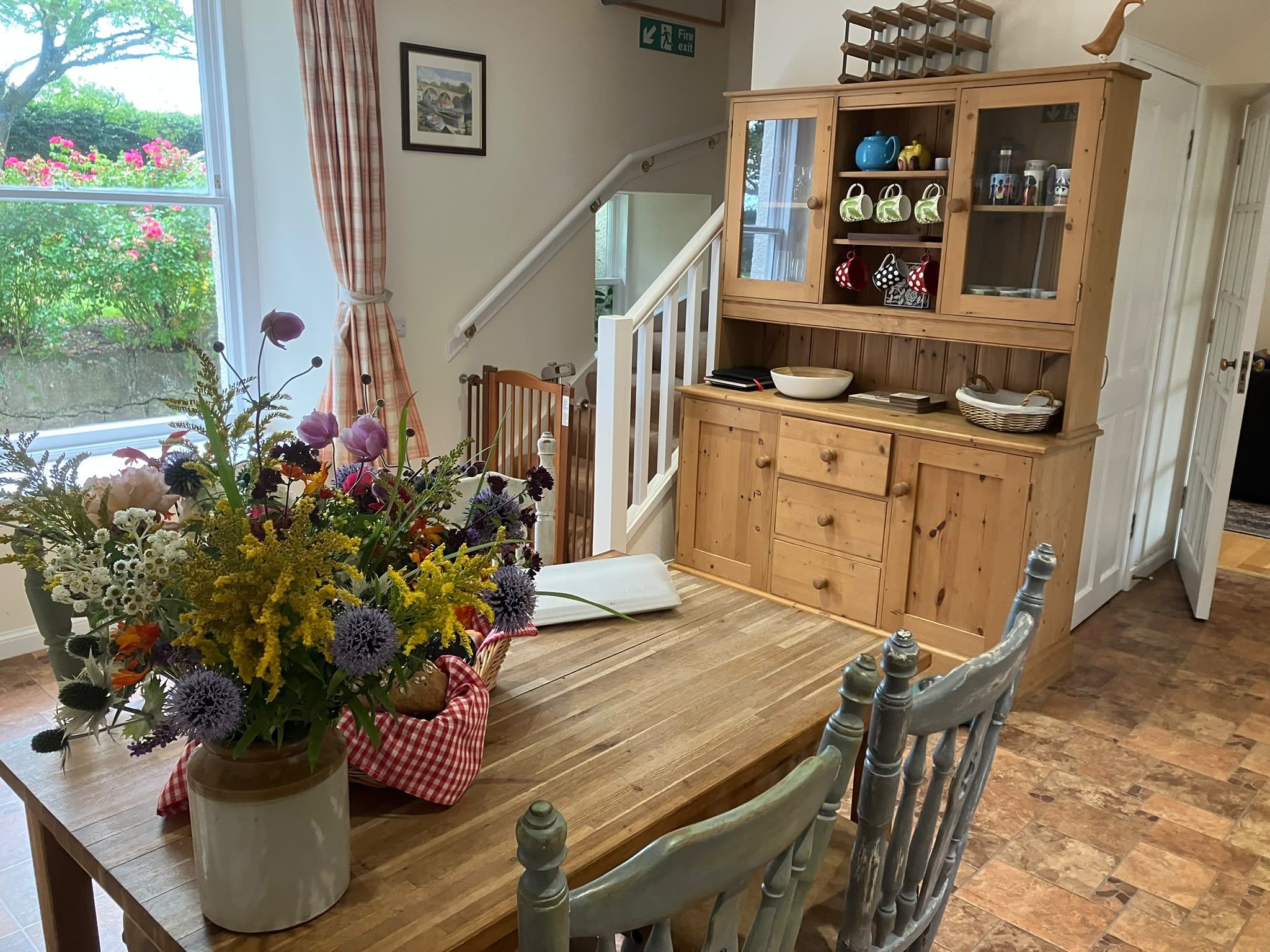 A dining area with a wooden table, chairs, a vase of flowers, a cabinet with kitchenware, and a staircase.