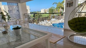 En balkon med et glasbord, hvide stole, en hængekøjsstol og udsigt til en swimmingpool og bygninger.
