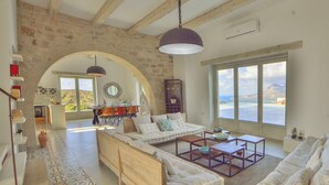 Ein geräumiges Wohnzimmer mit großem Sofa, Couchtisch und Essbereich. Eine Steinwand und der Blick auf das Meer durch große Fenster.