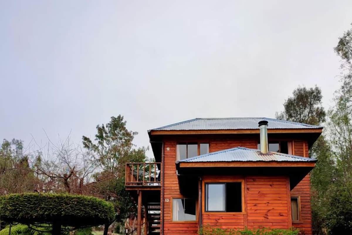 A two-story wooden cabin with a balcony, surrounded by a grassy yard and trees.