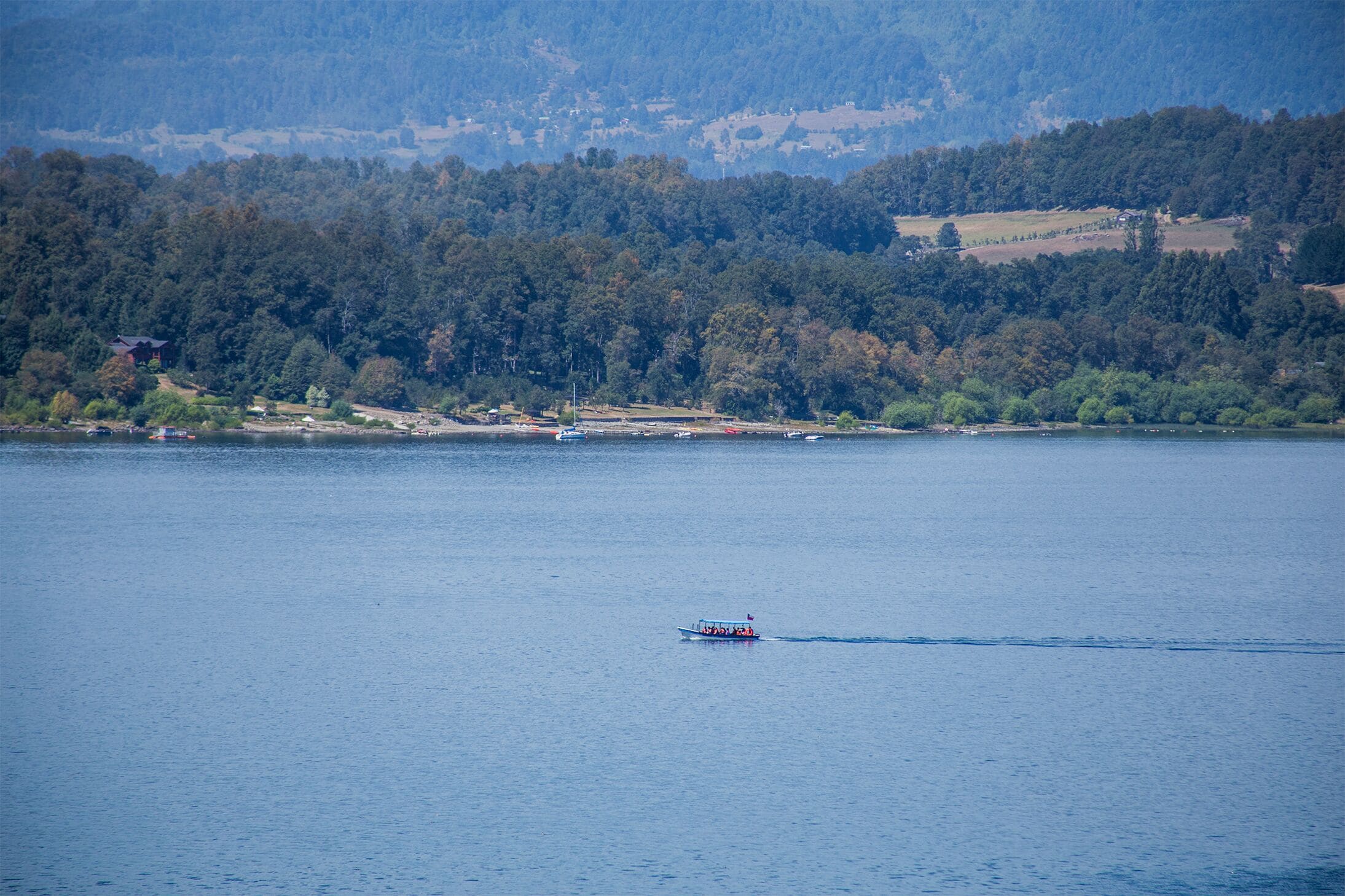 Ein See mit einem Boot im Vordergrund und einer bewaldeten Hügelkette im Hintergrund.