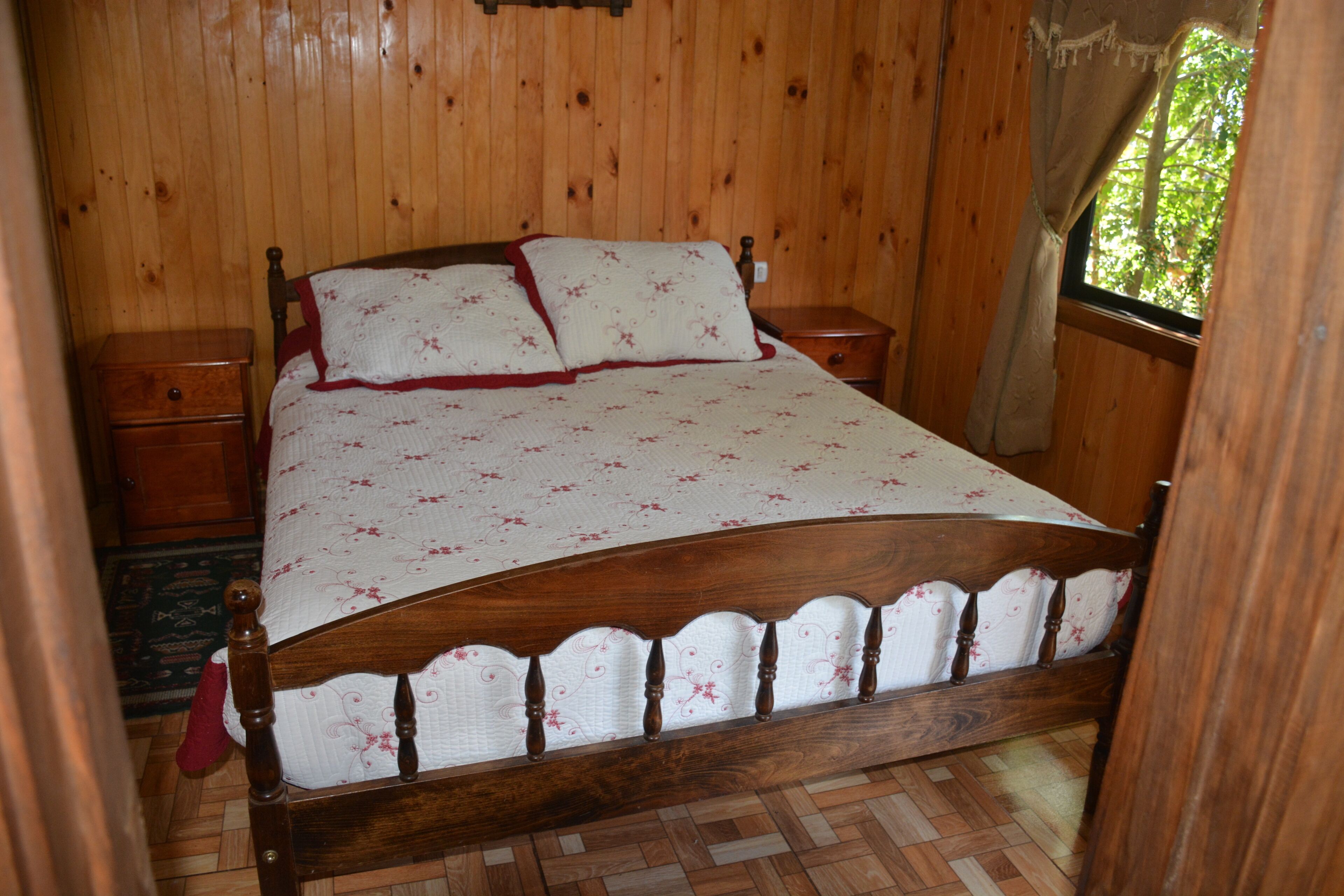 Un lit en bois avec un couvre-lit à motifs floraux et des oreillers, une table de chevet et une fenêtre donnant sur des arbres.