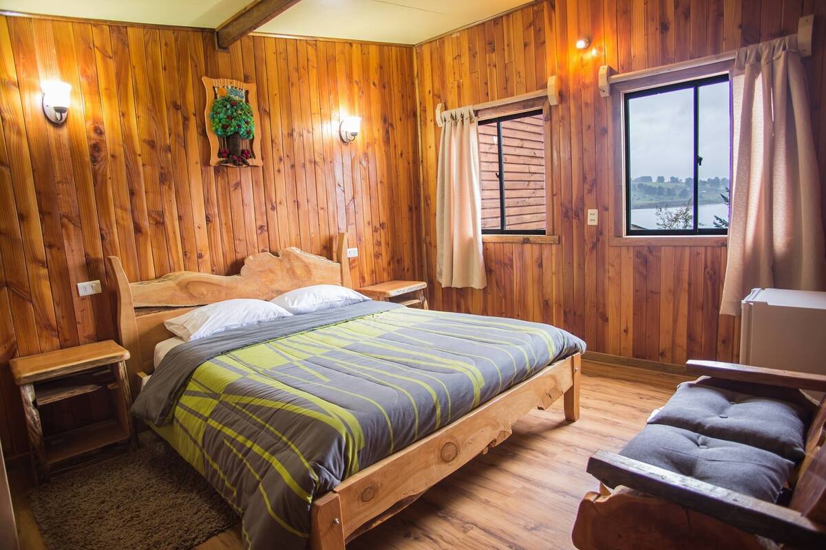 A wooden-paneled bedroom with a bed, bedside tables, a nightstand, a window with curtains, and a view of the outdoors.