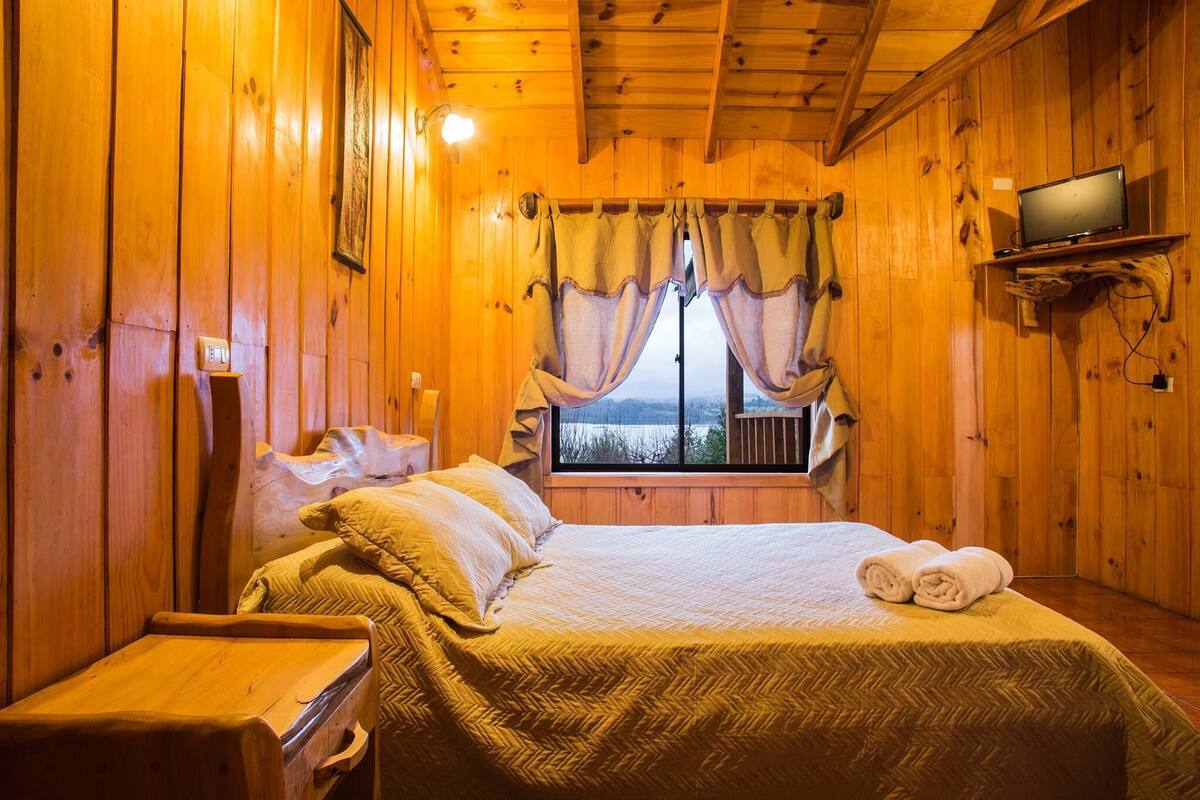 A wooden cabin room with a bed, a television on a shelf, and a window with curtains.