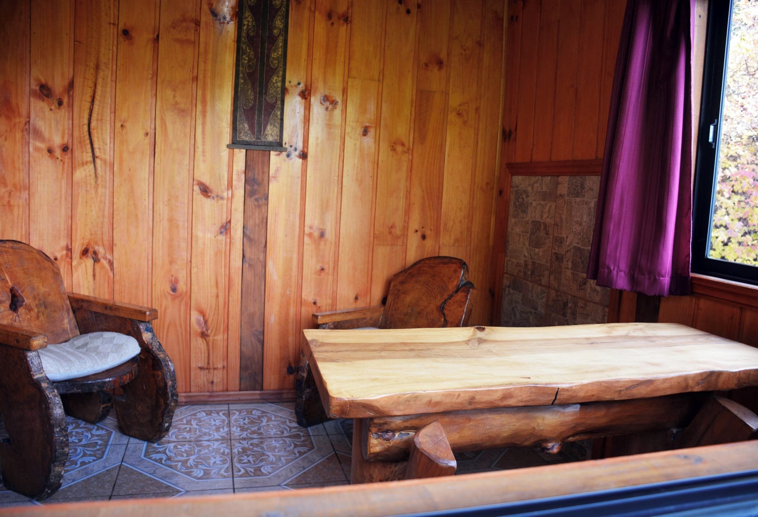 Un interior de madera con una mesa y dos sillas, una pared de piedra y una ventana con cortinas moradas.