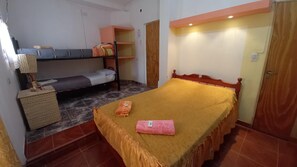 Una habitación con una cama individual, una cama doble, un armario de madera y una mesita con una lámpara.