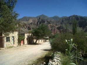 Un camino rural que conduce a un edificio de piedra con una puerta y una ventana, rodeado de árboles y montañas al fondo.