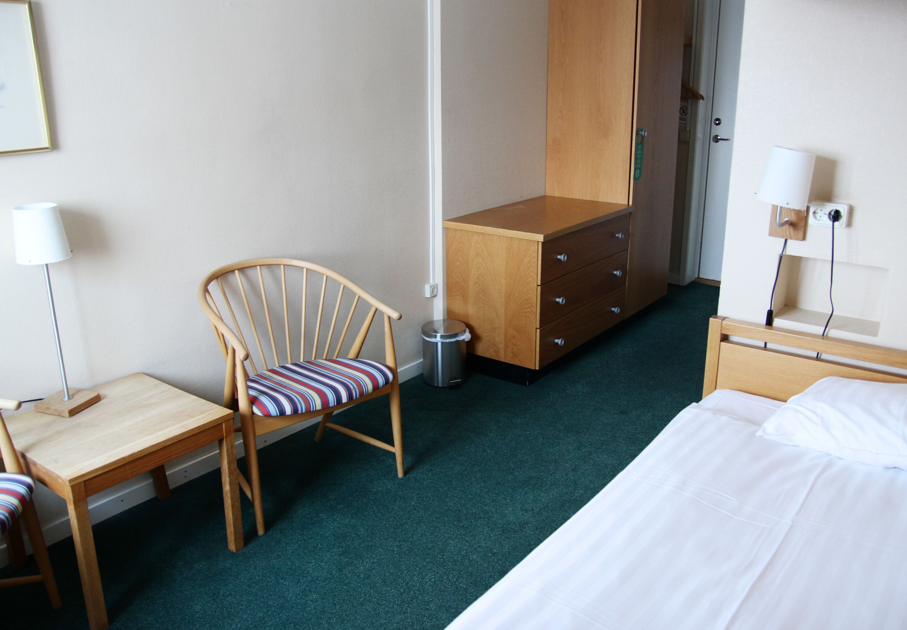 A hotel room with a bed, a wooden desk with a chair, a dresser, a trash can, and a lamp.