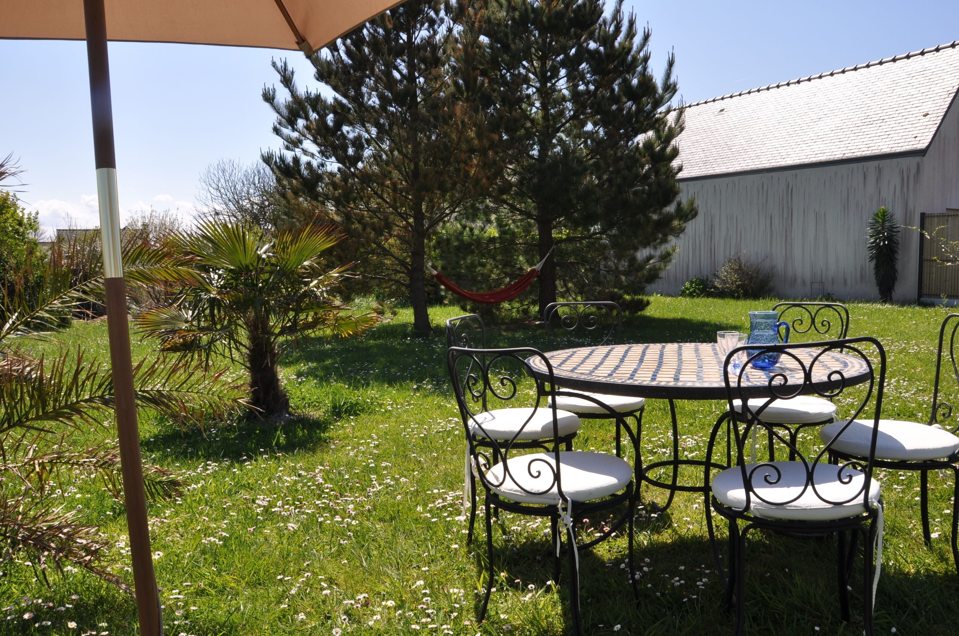 Un patio con una mesa redonda y cuatro sillas, rodeado de césped, árboles y una hamaca.
