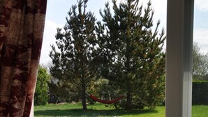 A view through a window with a curtain, revealing a grassy lawn with trees and a hammock. - Large, bright house with south-facing garden near the beach.  (Brignogan-Plage)