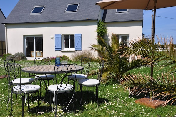 A house with a patio area featuring a round table and four chairs, surrounded by a garden with various plants and flowers.