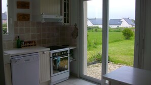 A kitchen with white cabinets, a stove, and a dishwasher. There is a window with a view of a grassy area and houses. - Large, bright house with south-facing garden near the beach.  (Brignogan-Plage)