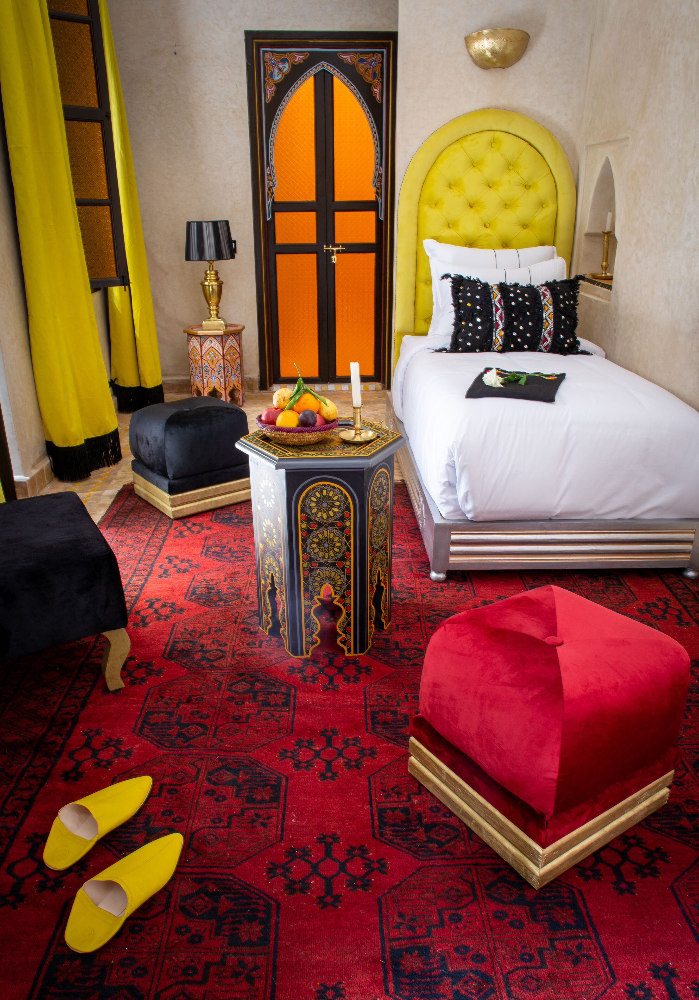 A room with a bed, a red ottoman, a small table with fruit, and a patterned carpet.