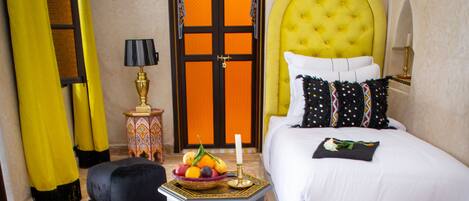 A room with a bed, a red ottoman, a small table with fruit, and a patterned carpet.