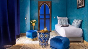 A room with a bed, a blue ottoman, a decorative table, and a door with intricate designs. - Riad Touda (Marrakech)