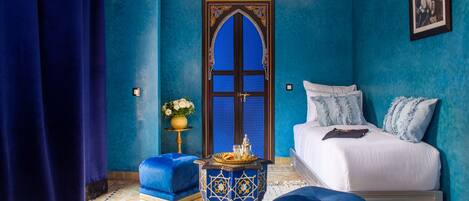 A room with a bed, a blue ottoman, a decorative table, and a door with intricate designs.