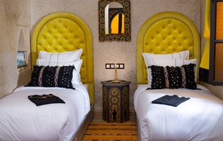 A hotel room with two beds, each with a tufted headboard, white bedding, and decorative pillows. There is a small side table with a lamp between the beds.