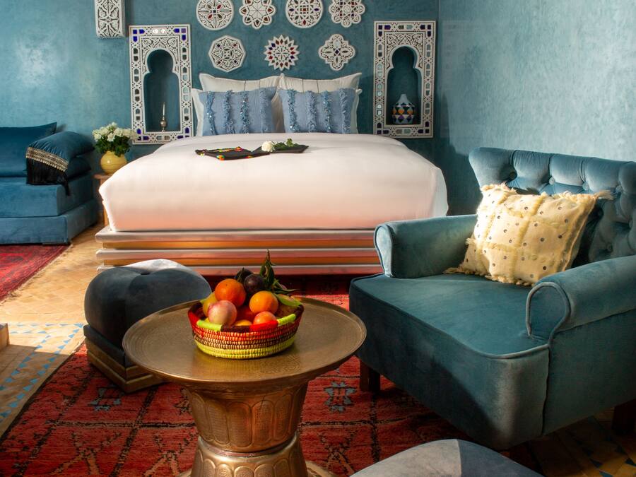 Un dormitorio con una cama grande, un sillón azul, una mesa redonda de centro con un frutero y una alfombra estampada.