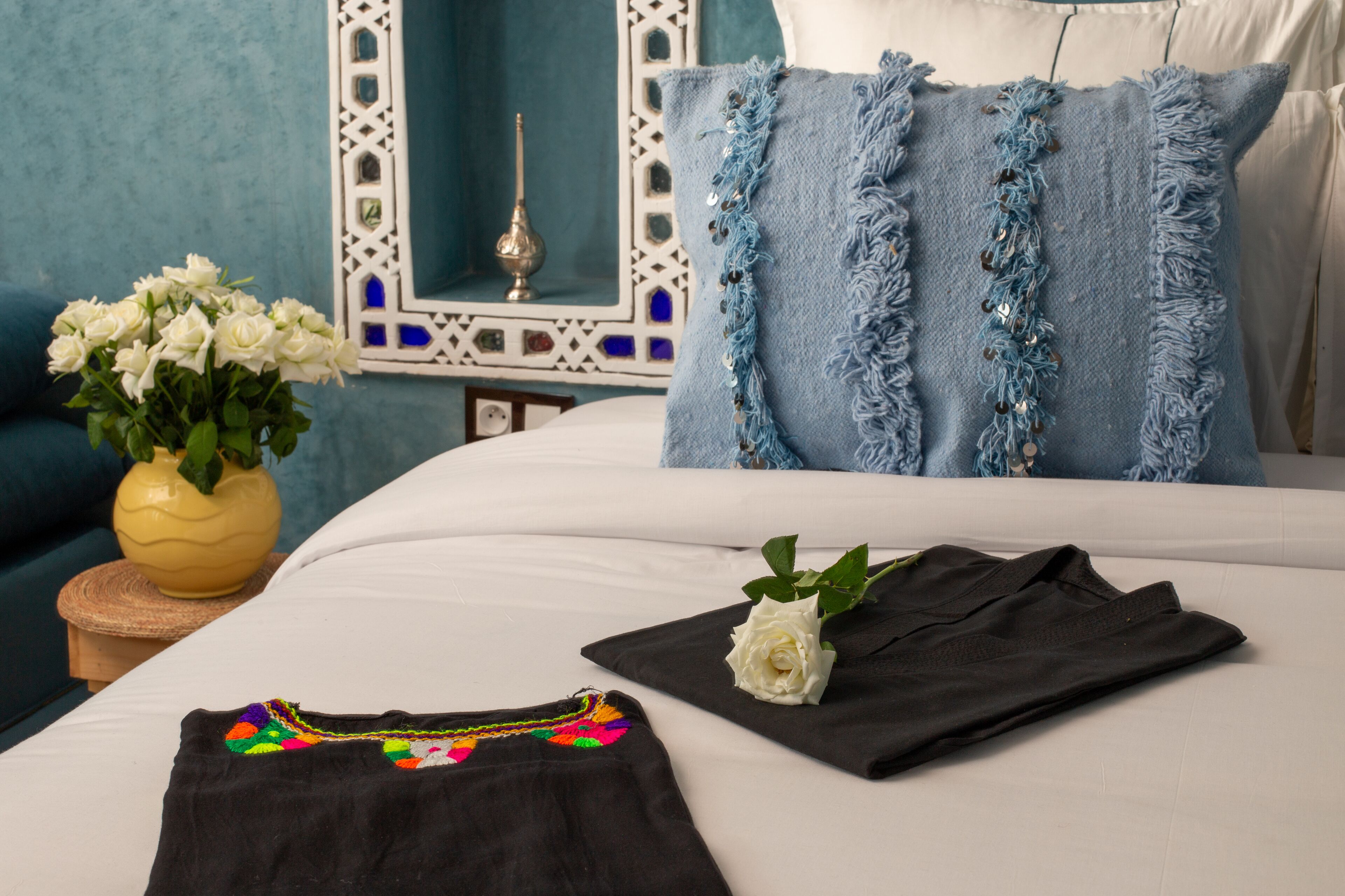 A neatly made bed with a white comforter and blue pillows, a black shirt with colorful embroidery, a white rose, and a vase of white flowers on a wooden nightstand.