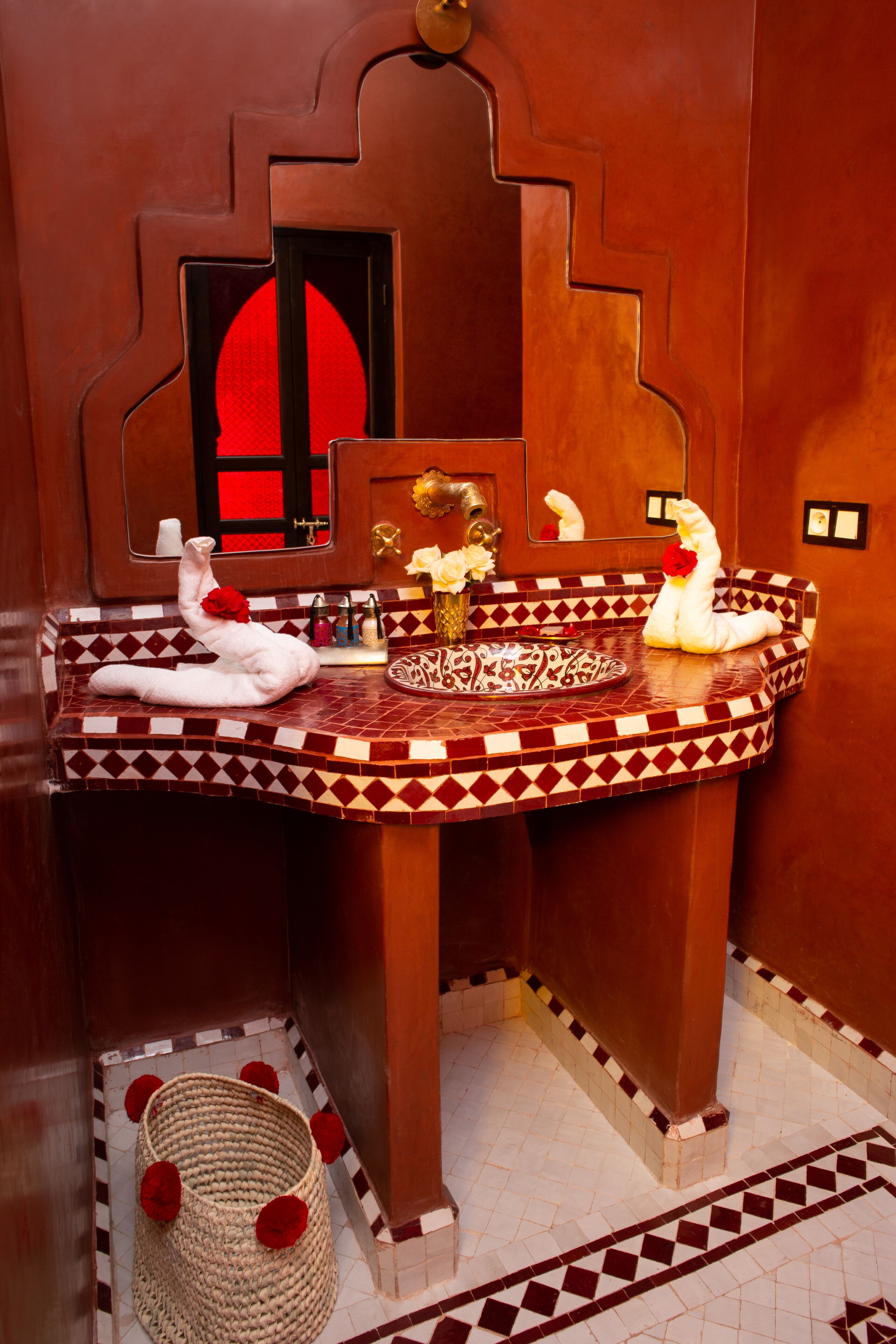 A bathroom with a red and white patterned tile design, a towel rack with a towel, a vase with flowers, and a decorative mirror.